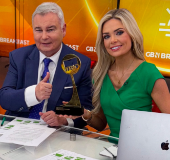 Two news anchors at a desk, one holding an award and giving a thumbs up.