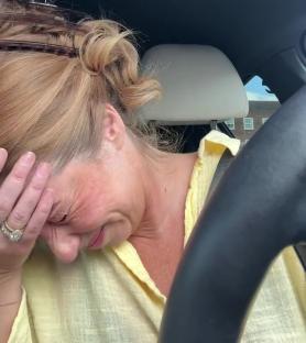 A woman sits in a car, crying and holding her head.