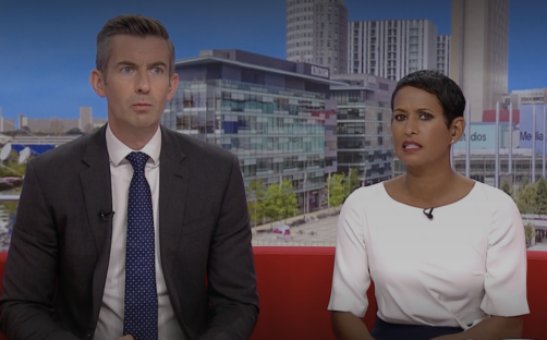 Screenshot of two news anchors sitting on a red couch.