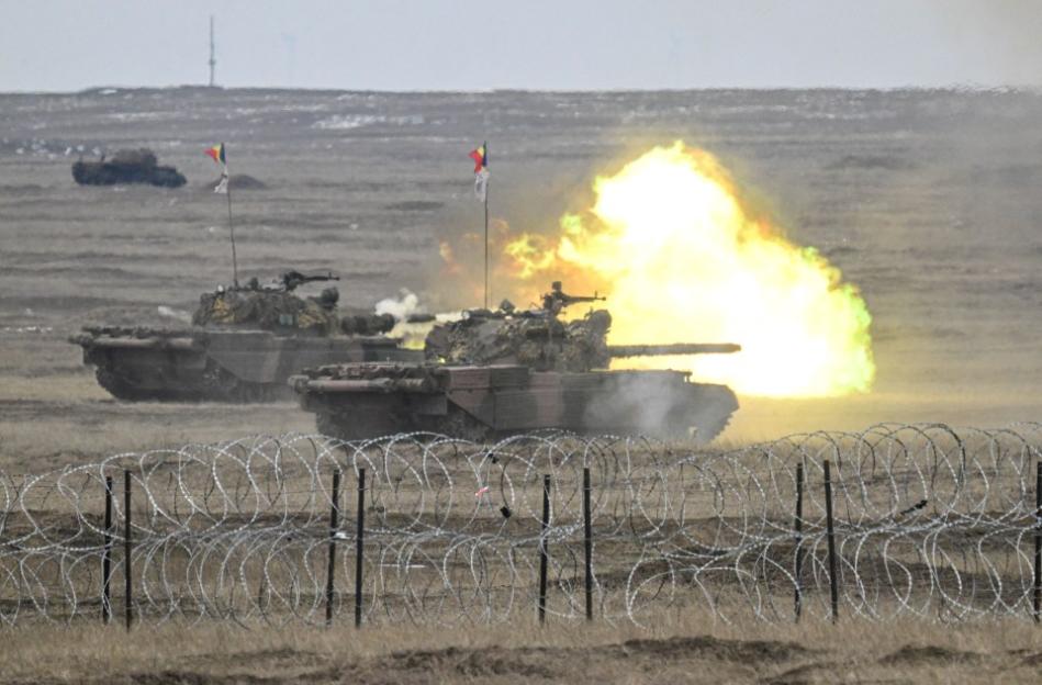 Two tanks firing behind a barbed wire fence.