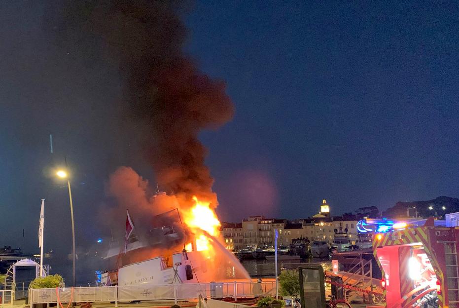 A superyacht engulfed in flames in Saint-Tropez harbor, with firefighters battling the blaze.