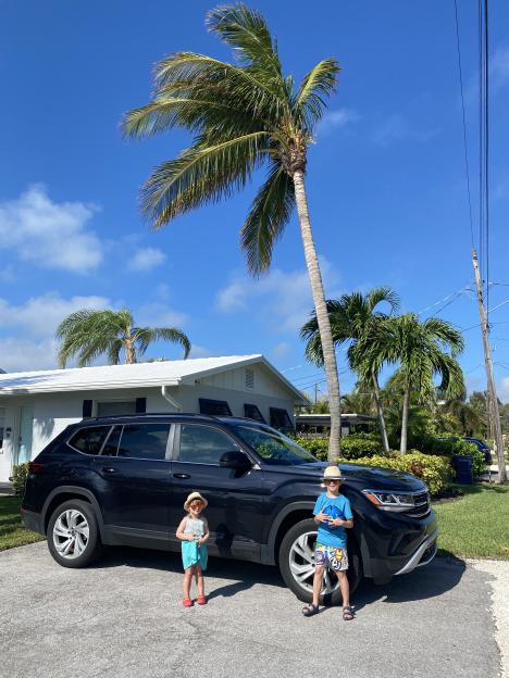 Two children standing by a dark-colored SUV in front of a house with palm trees.