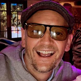 Close-up portrait of a smiling man wearing sunglasses and a cap.