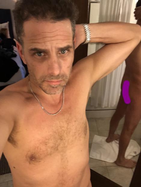 Man with chest hair and necklace, reflected in mirror with another person's legs visible.