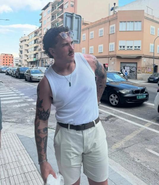 A man in a white tank top and beige shorts stands on a city street.
