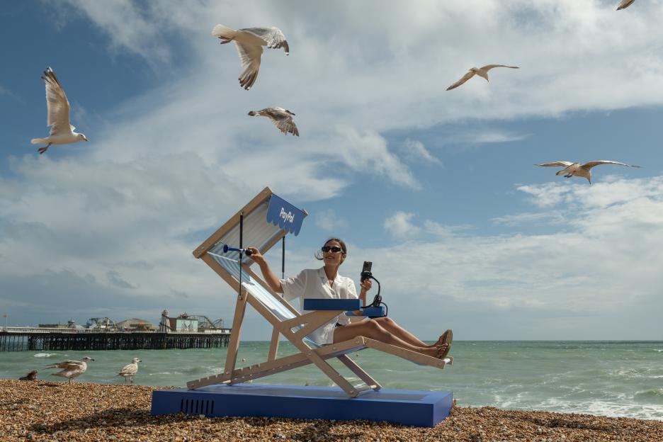 Woman using phone in a PayPal branded deck chair on the beach.