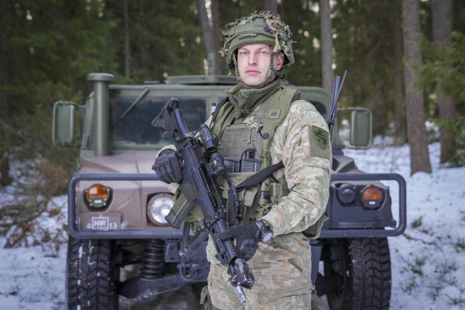 Lithuanian soldier in winter camouflage holding a rifle in front of an army vehicle.