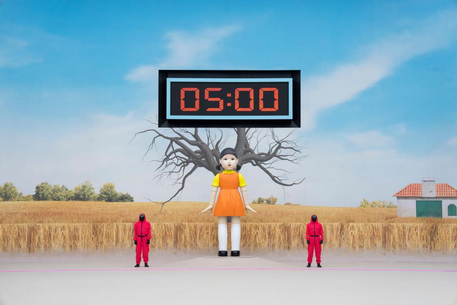 Squid Game: The Challenge set with a large doll, countdown timer, and masked figures.