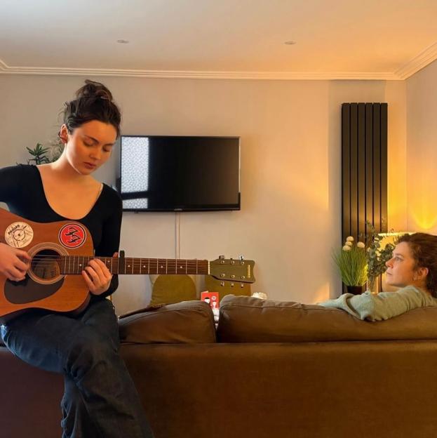 Gracie playing guitar in living room with mum Anna relaxing on sofa.