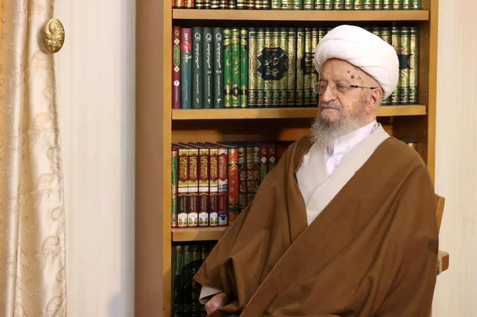 Man in brown robe sitting in front of a bookshelf.