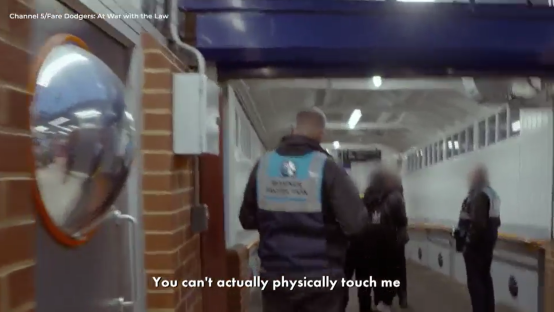 Screenshot of a security guard and other people walking down a corridor.
