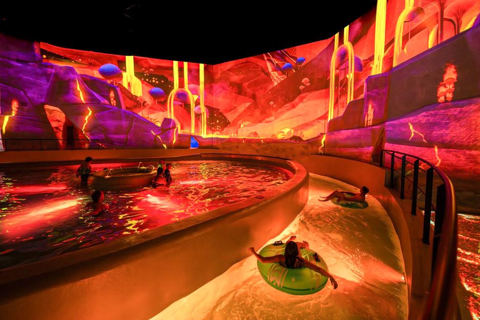 People in inner tubes floating down a waterpark's lazy river with a vibrant, projected landscape.