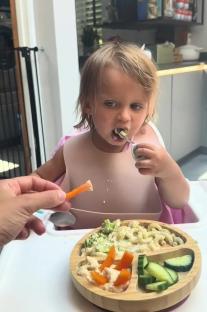 Toddler eating a meal of pasta, broccoli, hummus, carrots, and cucumbers.