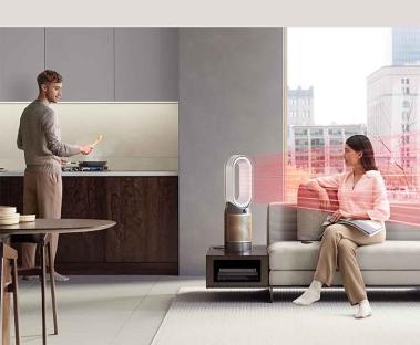 Person cooking in a kitchen while another person relaxes on a couch warmed by a Dyson Pure Hot+Cool purifier.