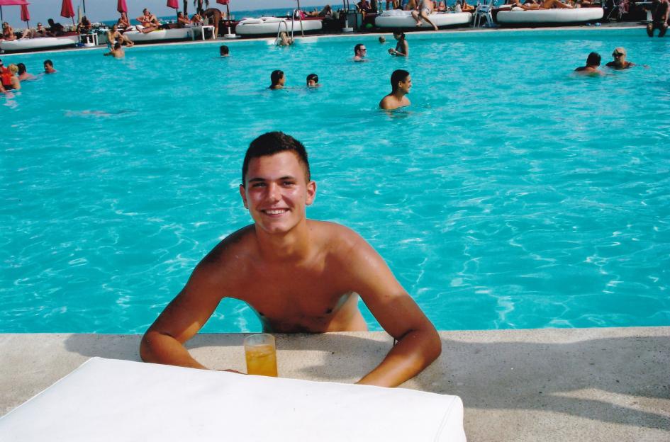 A young man sits at the edge of a swimming pool with a drink.