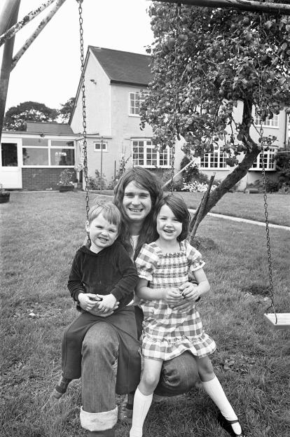 Black and white photo of Ozzy Osbourne with his children on a swing set in his backyard.