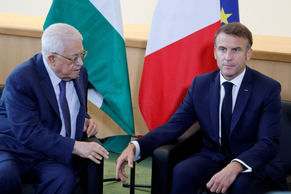 Two men in suits sit and talk, with flags of Algeria and France behind them.