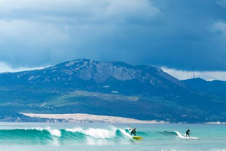 Two people paddleboarding on ocean waves with a mountain in the background.