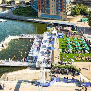 Aerial view of a summer splash event at Royal Docks, featuring a floating pool, sunbathers, and a beach area.