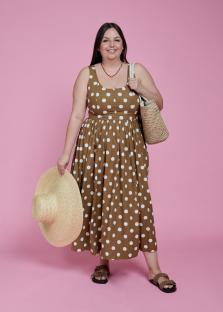 Woman in brown polka dot dress holding a straw hat and bag.