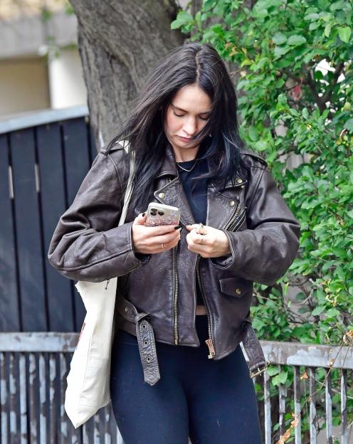 Lily James with dark hair, wearing a brown leather jacket, looking at her phone.