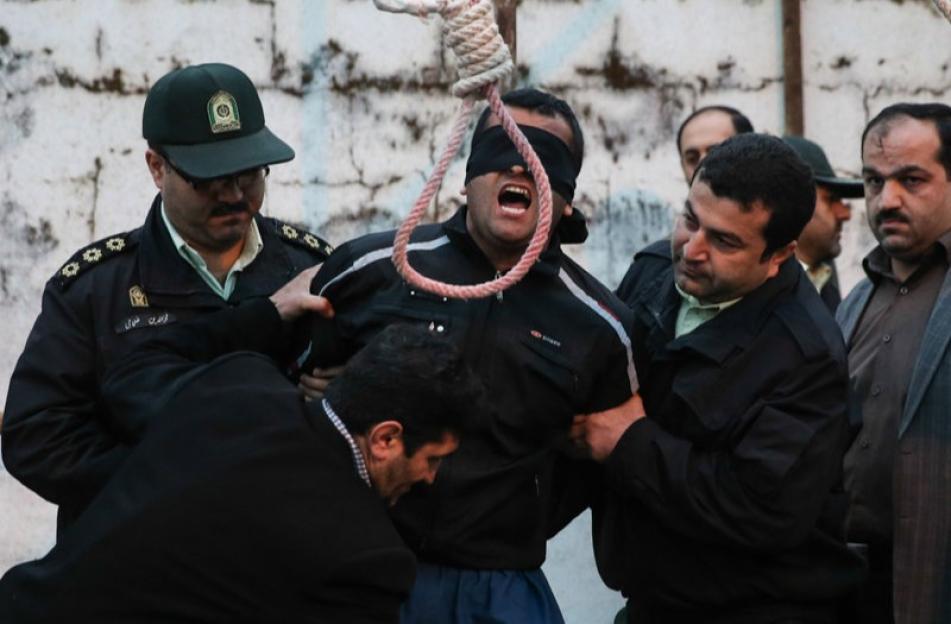 A blindfolded man about to be hanged is held by law enforcement officials; the victim's family forgave him.
