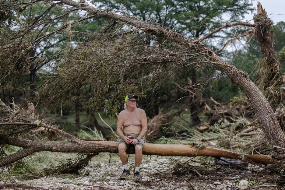 Man sits on a fallen tree trunk amidst flood debris.