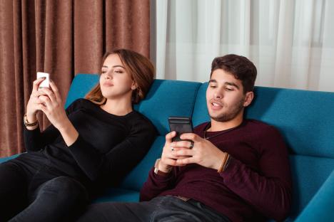 A young couple sits on a couch, each using a cellphone.