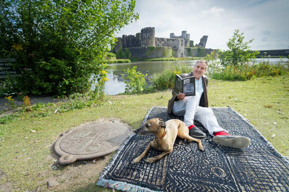 Man reading a book about manhole covers with his dog near a castle.