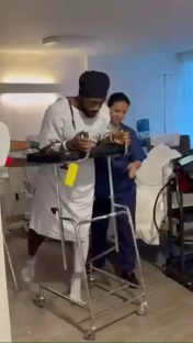 Man in hospital gown using a walker with assistance from a nurse.