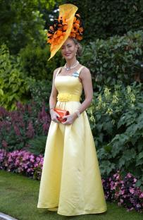 Woman in yellow dress and large orange and black hat at a racecourse.