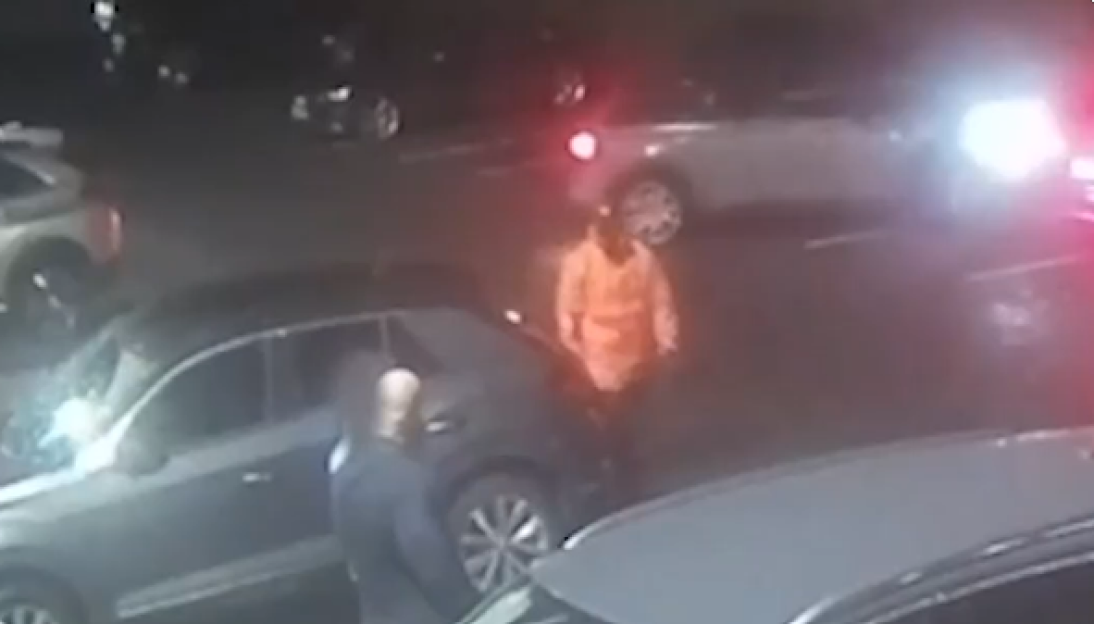 Nighttime security footage of a person in an orange jacket near several cars.