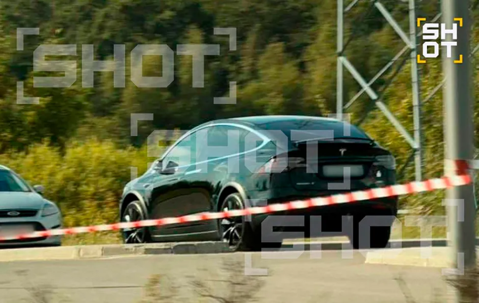 A dark-colored Tesla Model X driving past a red and white striped barrier.