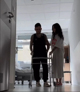 Man using a walker in a hospital hallway, assisted by a woman.