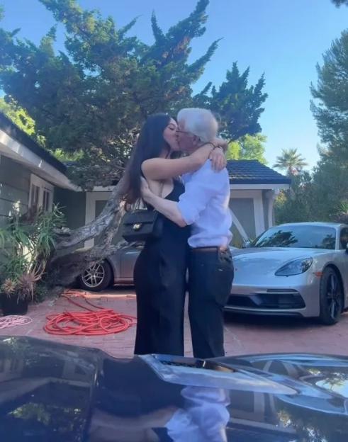 A young woman and an older man kissing in front of a house and cars.