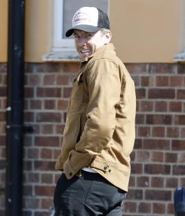 Danny Jones of McFly, wearing a tan jacket and baseball cap, smiles.