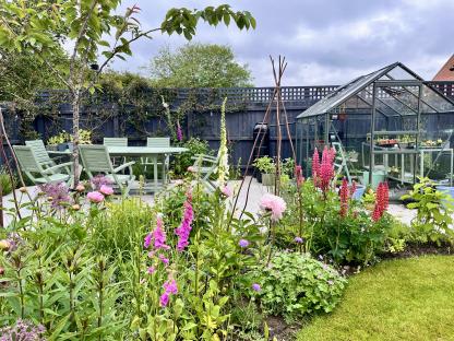 A spring garden with blooming flowerbeds, a patio table and chairs, and a greenhouse.