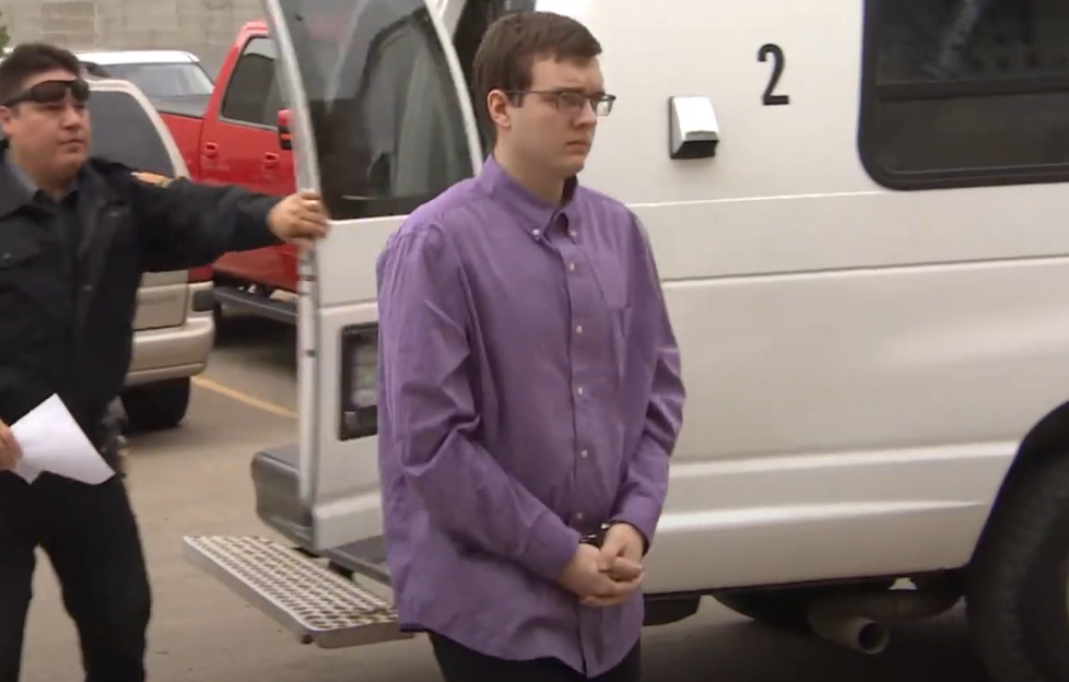 A handcuffed man in a purple shirt being escorted by a police officer.