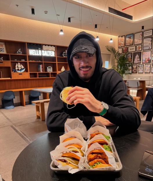 Man holding a burger, sitting at a table with several more burgers in front of him.