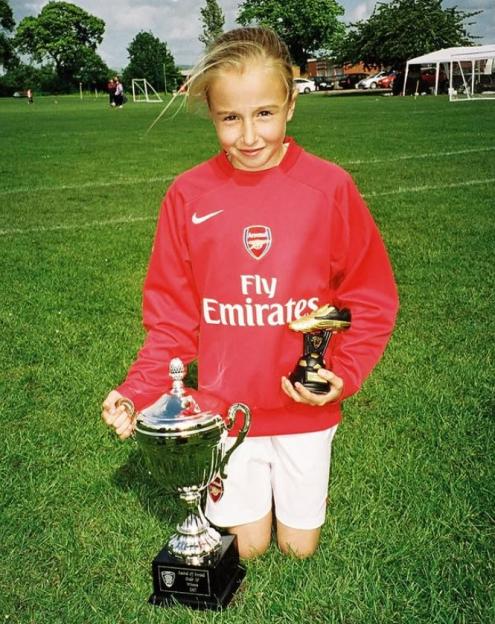 Girl in Arsenal jersey kneeling on grass, holding a trophy and a golden boot award.