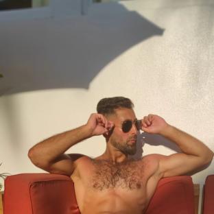 Shirtless man relaxing on a red couch, adjusting his sunglasses.
