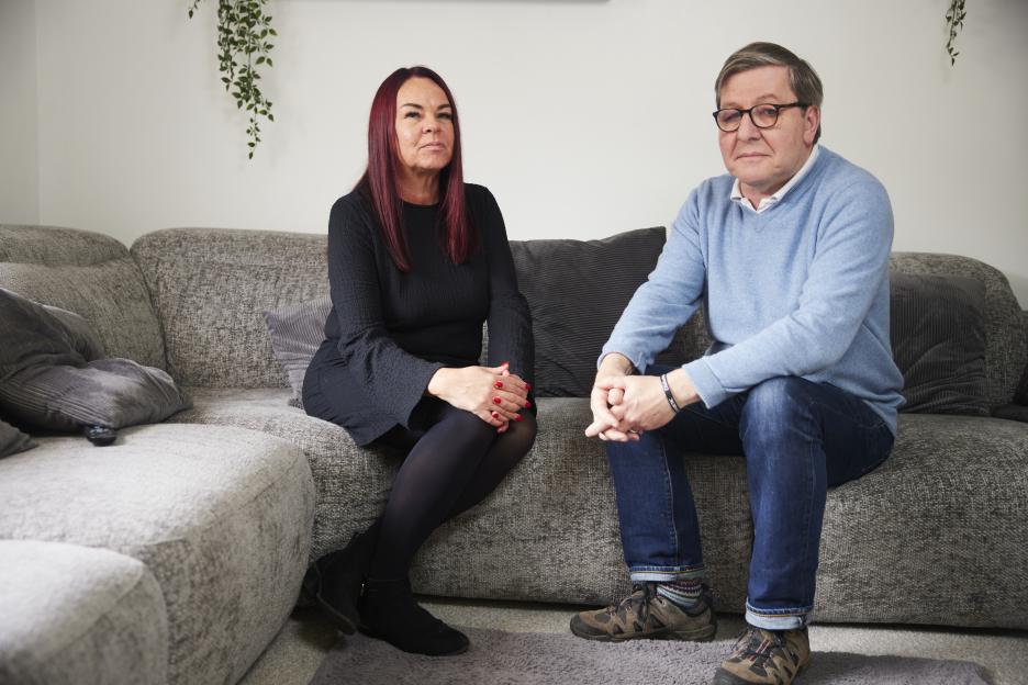 A woman and man sit on a couch, looking at the camera.