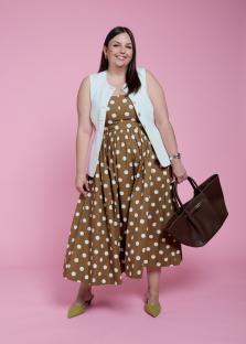 Woman in brown polka dot dress and white vest holding a brown handbag.