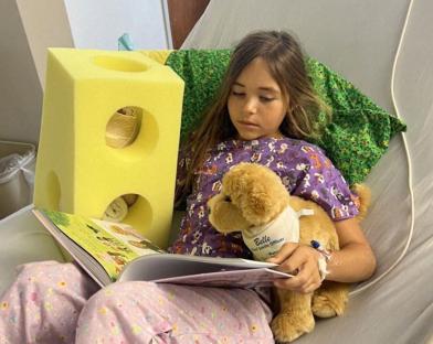 Girl in hospital bed reading book with stuffed dog.