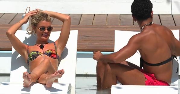 Woman in bikini and sunglasses sits on a lounge chair by the pool talking to a man in red swim shorts.