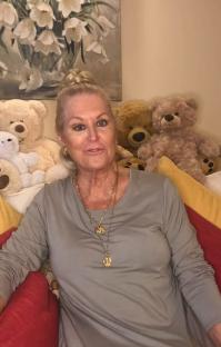 Woman with blonde hair wearing a gray shirt and necklaces sits on a couch surrounded by teddy bears.