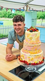 Man smiling proudly next to a three-tiered cake.