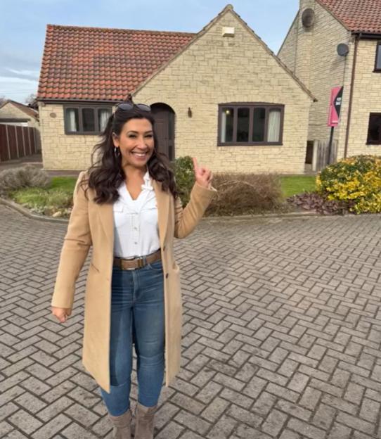A woman stands in front of two houses; a large stone house and a smaller, more modern house.