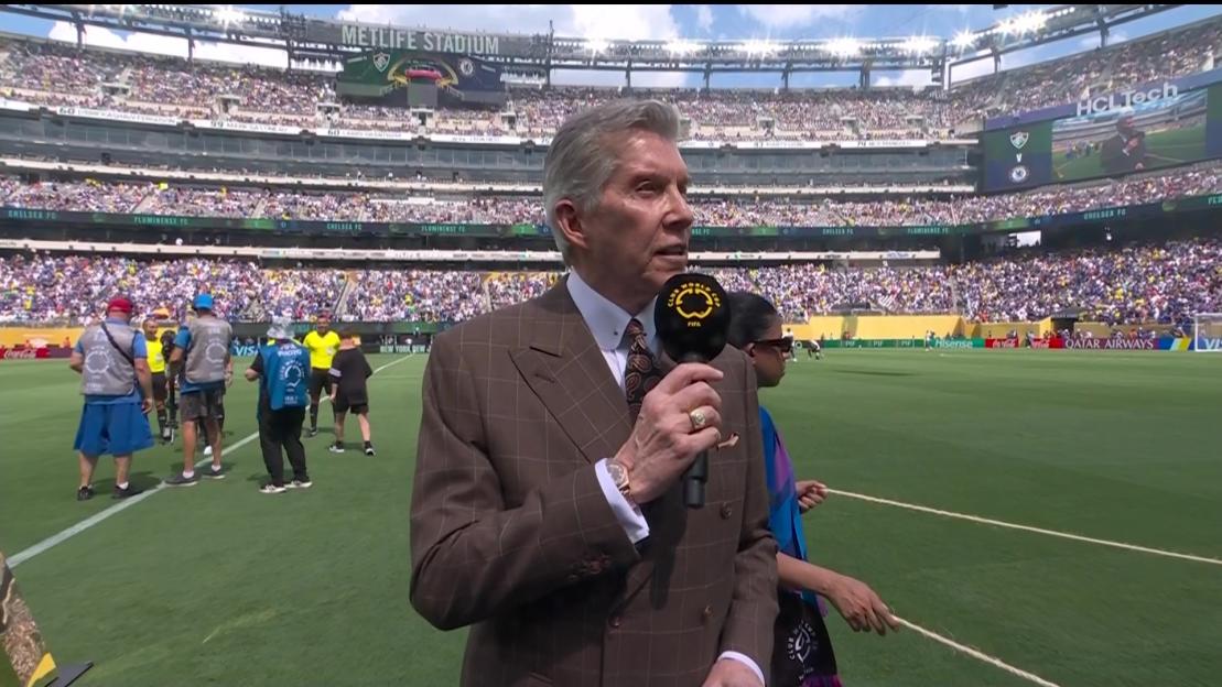 A man in a suit conducts a televised interview at a full soccer stadium.