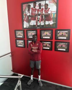 Man in Arsenal attire standing in front of framed photos of Arsenal youth teams.